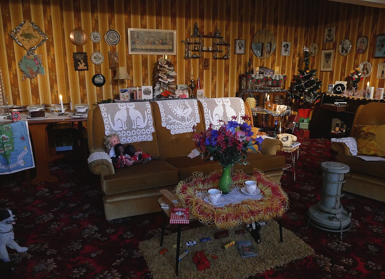 A museum room display with patterned furnishings, crochet, ornaments and a glass cabinet.