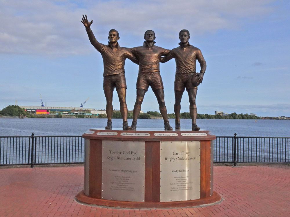 Three bronze rugby players stand with their arms around one another on the Rugby Codebreakers monument in Cardiff Bay.