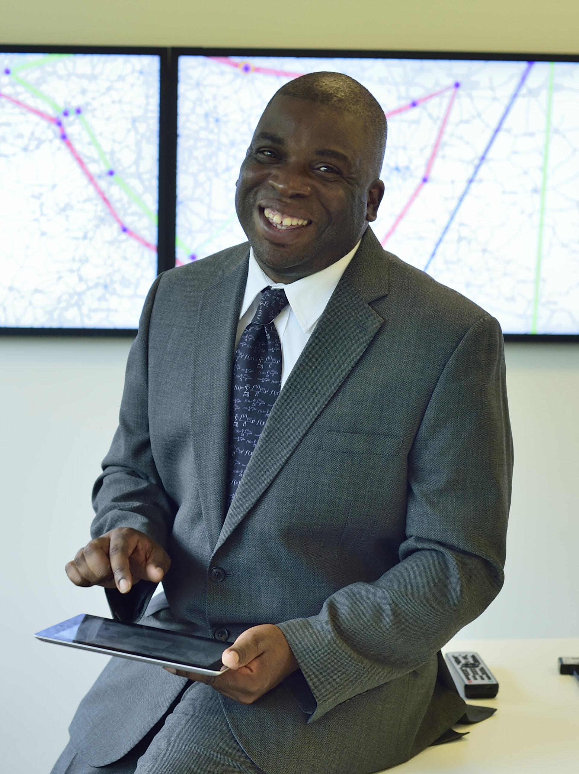 Nira Chamberlain smiling in a grey suit and a tie patterned with mathematical symbols, holding a tablet.