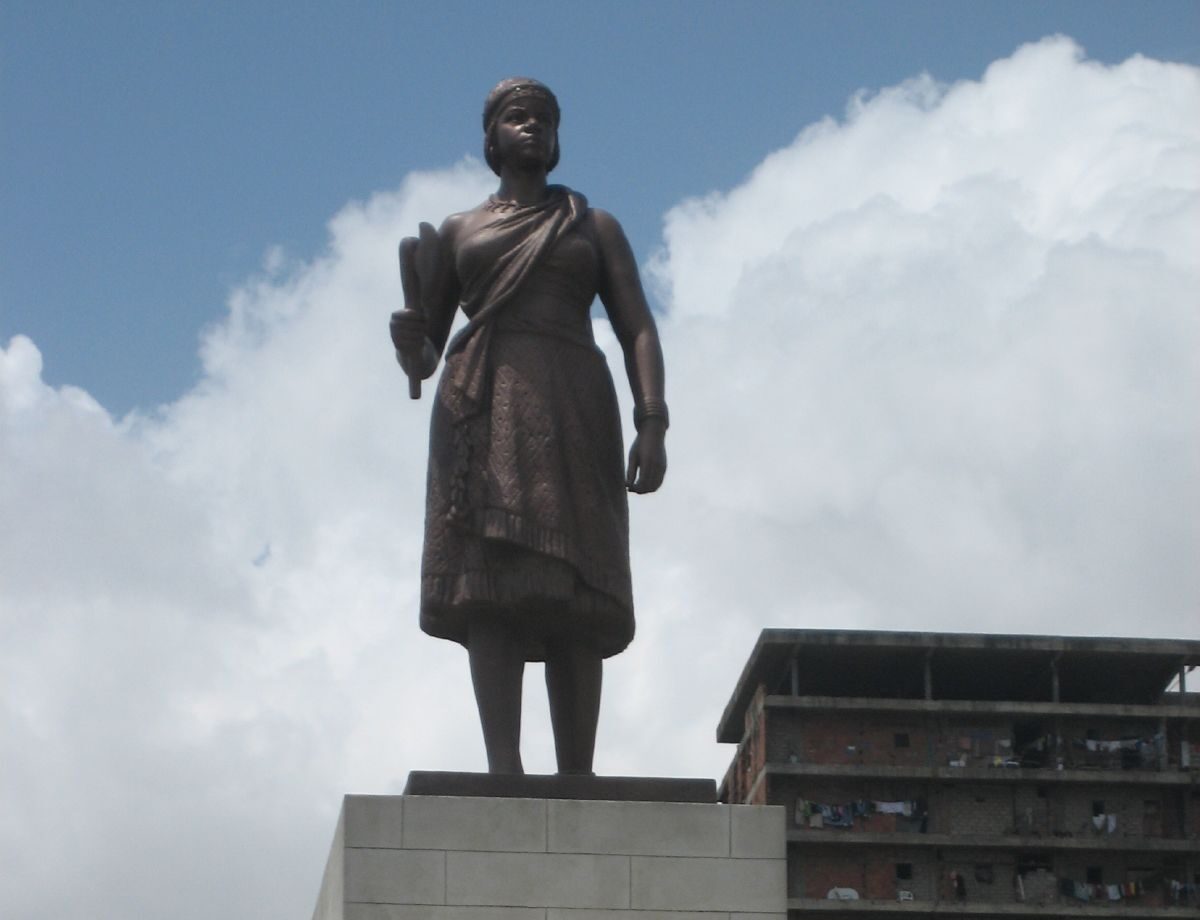 A statue of Queen Njinga holding an axe stands on a tall stone plinth against a cloudy sky in Luanda.