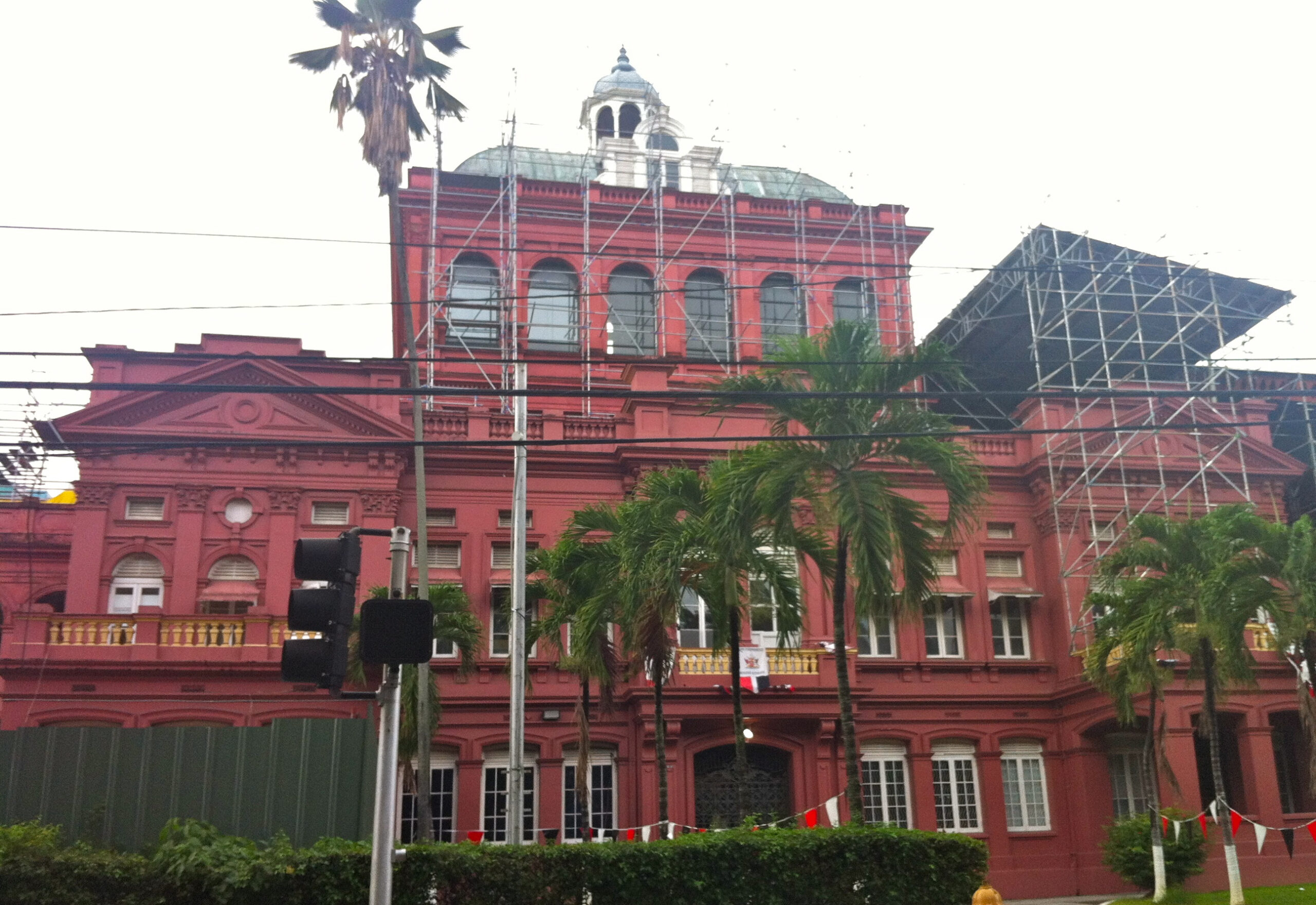 The red facade of the Red House in Port of Spain, with scaffolding around the central tower and palm trees in front.