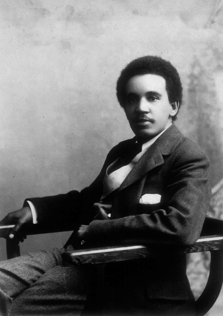 A monochrome studio portrait of Samuel Coleridge-Taylor seated in a wooden armchair, wearing a dark suit and a light waistcoat.